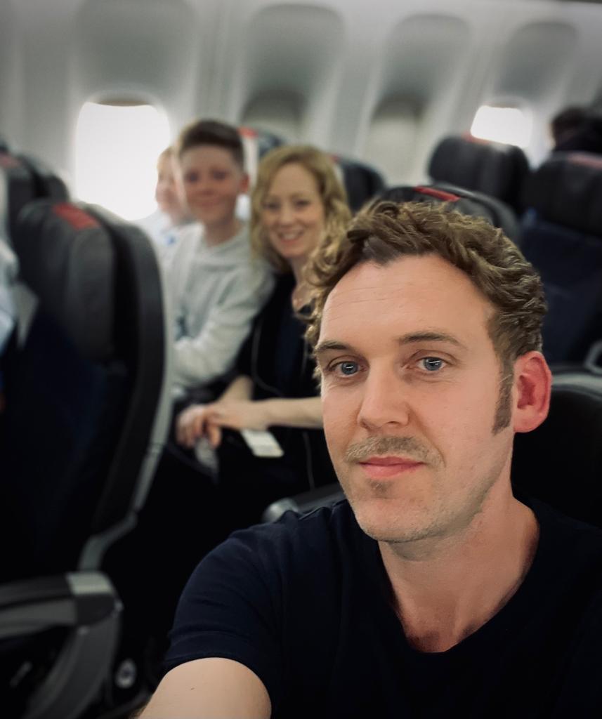 A selfie picture of the thompson family taken by Rory, sitting on a plane. Rory is inm focus and the family are blurred