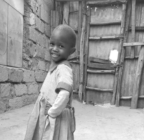 A picture of a young girl from kenya smiling with her hands in her dress pockets. 