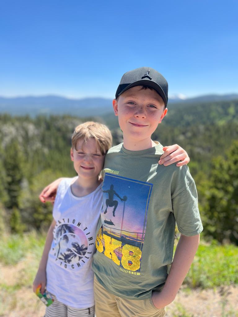 Two young boys standing next to each other with the arms around each other smiling with mountains behind them