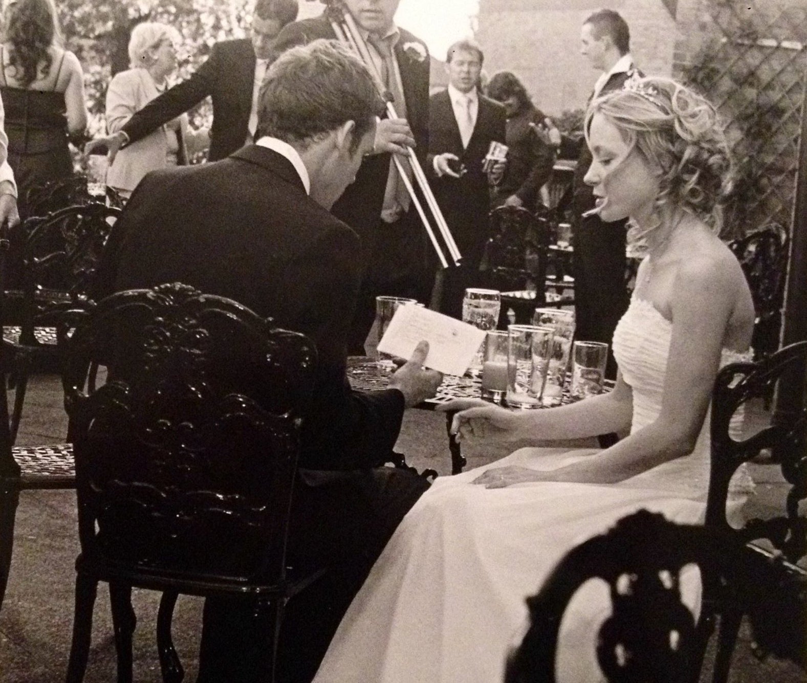 Rory and Sarah on their wedding day. They are sitting outside surrounded by guests. Rory's back is toward the camera and he is wearing a black suit and which shirt. Sarah is wearing a strapless white wedding dress, sitting to rory's right. 