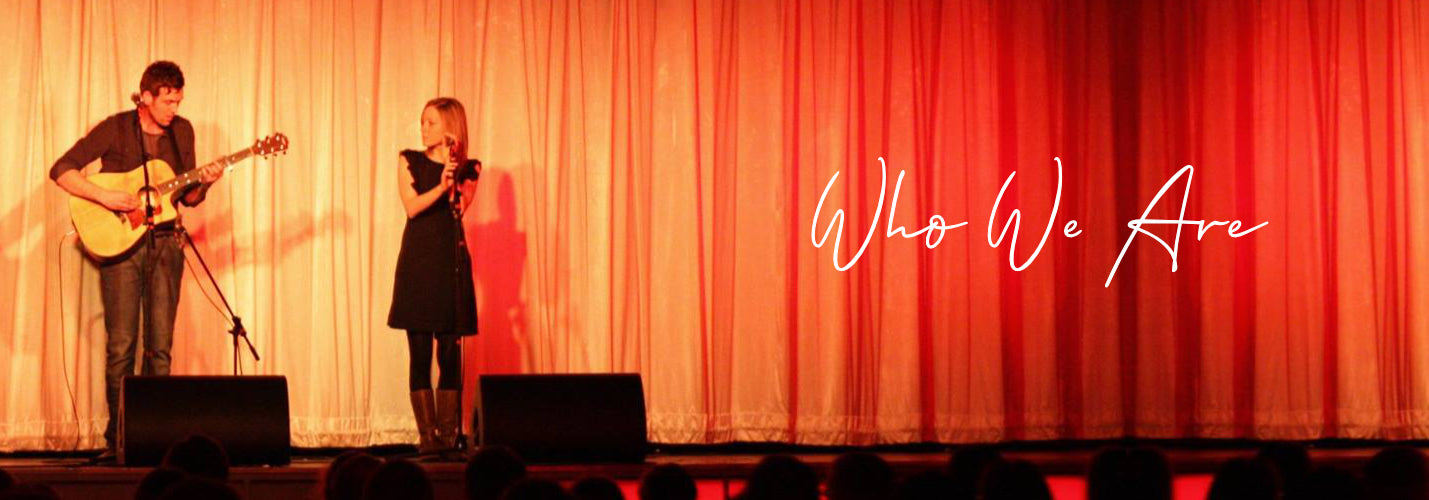 Rory and Sarah performing on a stage, Rory holding a guitar and Sarah standing by a microphone. There is an orange glow across the stage curtains which are closed. The words 'Who We Are' are also on the image