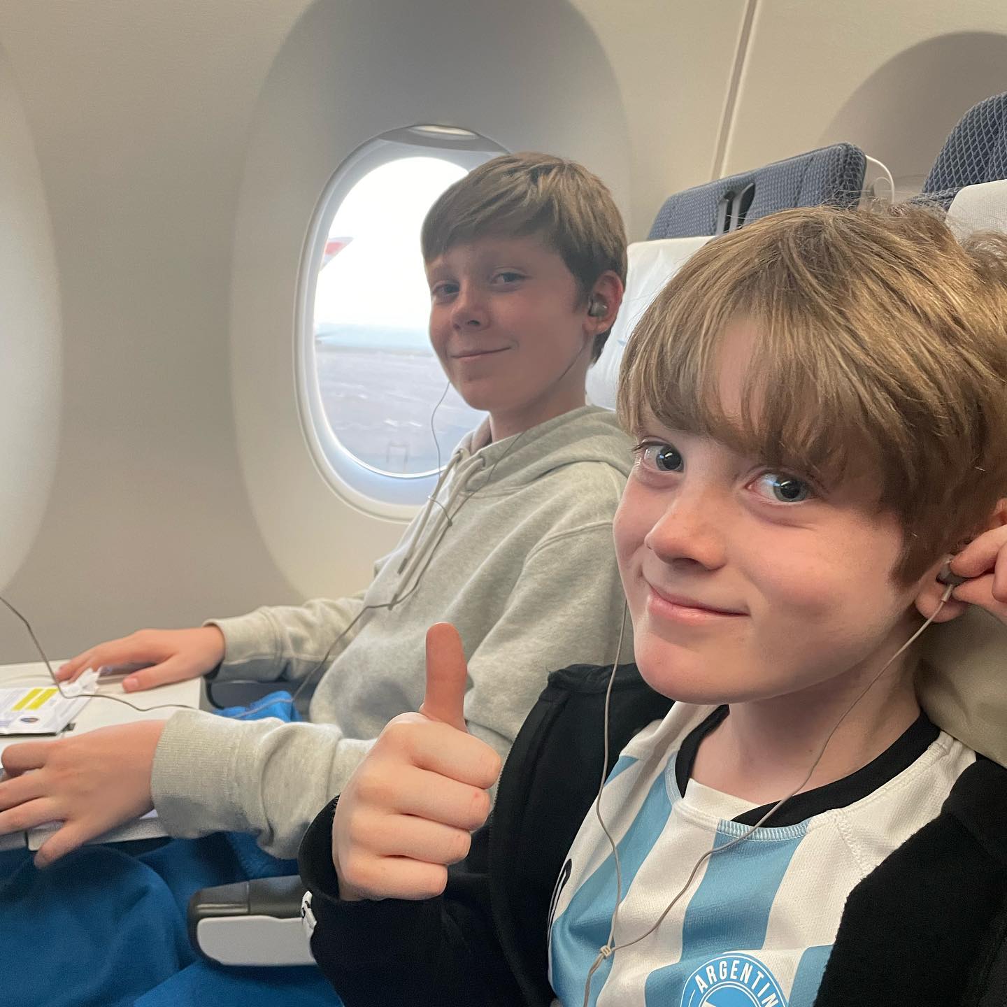 Fynn and Oscar sitting on a plane, Fynn next to the window smiling, Oscar with his thumb up smiling