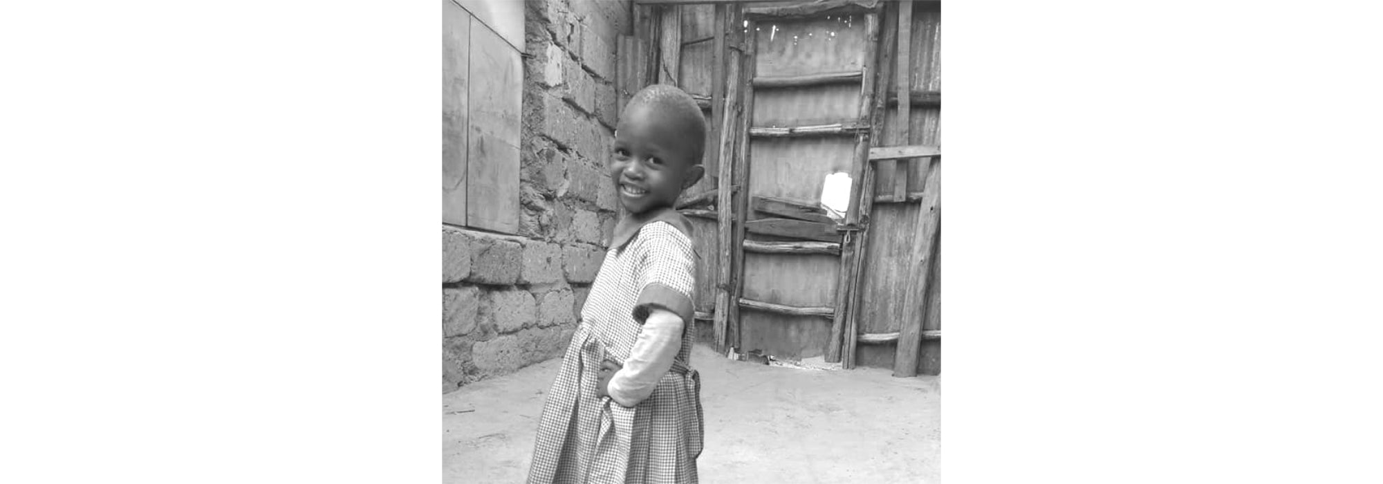 A picture of a young girl from kenya smiling with her hands in her dress pockets. 