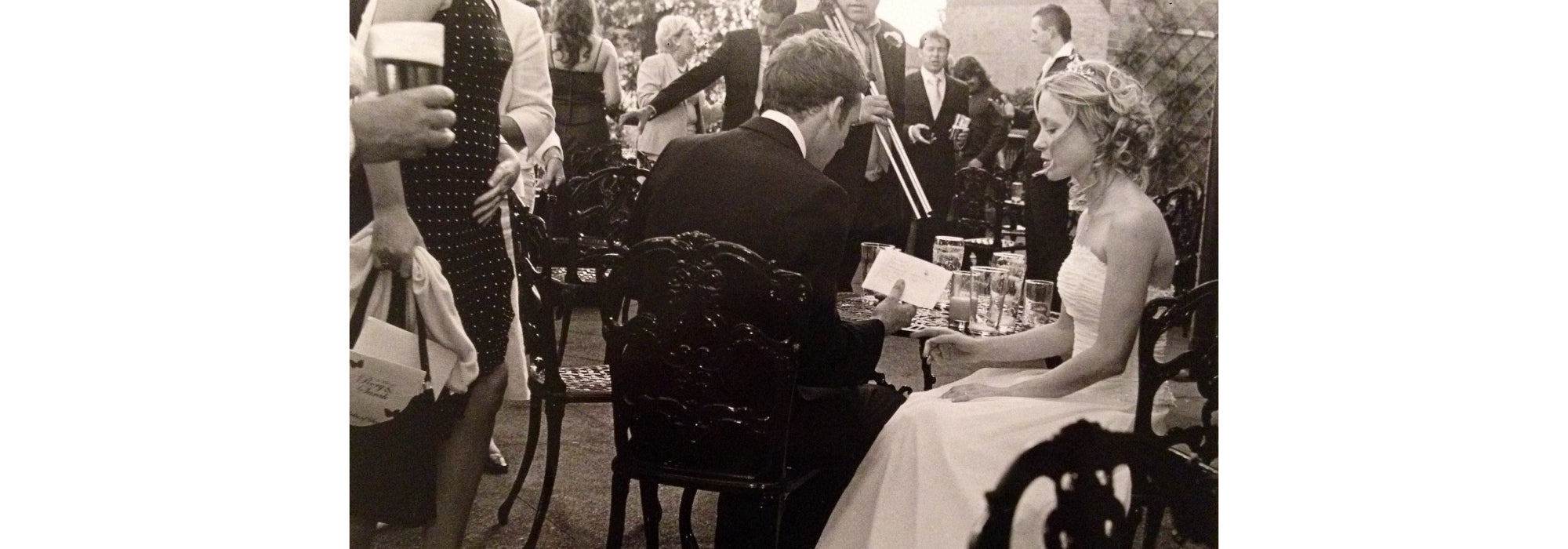 Rory and Sarah on their wedding day. They are sitting outside surrounded by guests. Rory's back is toward the camera and he is wearing a black suit and which shirt. Sarah is wearing a strapless white wedding dress, sitting to rory's right. 