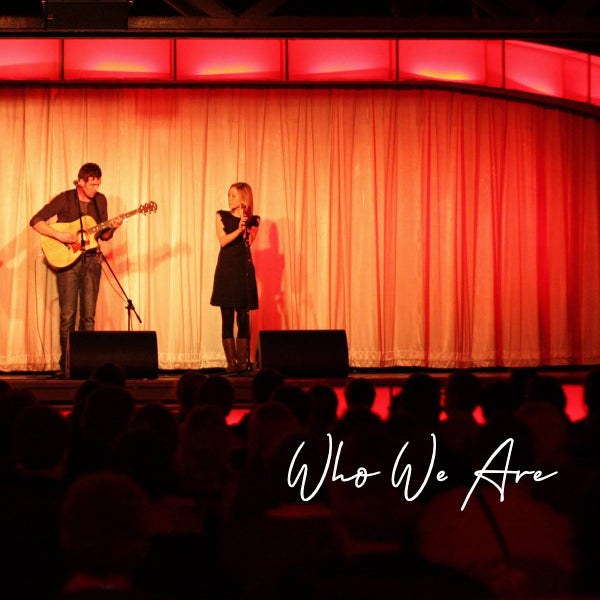 Rory and Sarah performing on a stage, Rory holding a guitar and Sarah standing by a microphone. There is an orange glow across the stage curtains which are closed. The words 'Who We Are' are also on the image